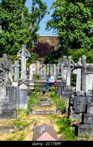 Man sitting on a bench, reading newspaper, Brompton Cemetery, London, UK Stock Photo