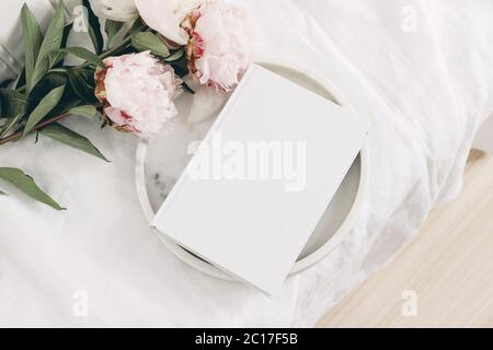 Wedding still life scene. White empty book cover mockup on marble tray. Pink peony flowers on white linen table cloth. Vintage feminine styled photo Stock Photo