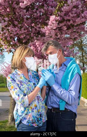 Elderly loving couple wearing surgical masks and gloves standing close together holding hands under a tree in a street covered in colorful pink spring