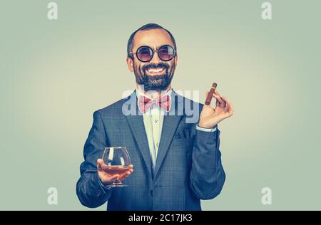 The smiling bearded man in formal wear and necktie is smoking cigar holding whiskey glass alcohol looking at camera happy isolated on green yellow uni Stock Photo