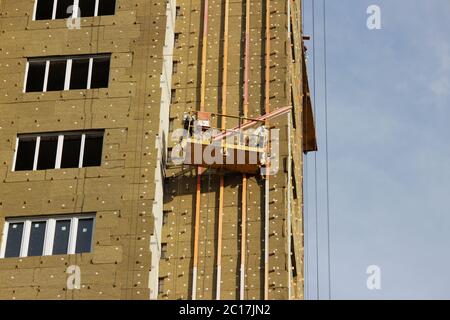 worker in yellow suspended cradle mounts environmental boards for insulation on a newly built high-rise building Stock Photo