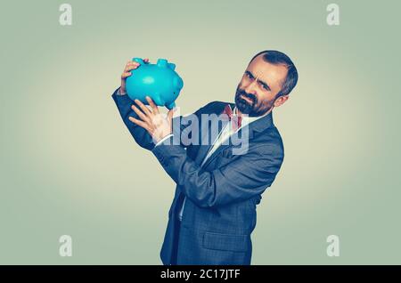 Adult displeased bearded man in elegant suit with red bow tie holding overturned piggy bank, showing its emptiness. Mixed race bearded model isolated Stock Photo