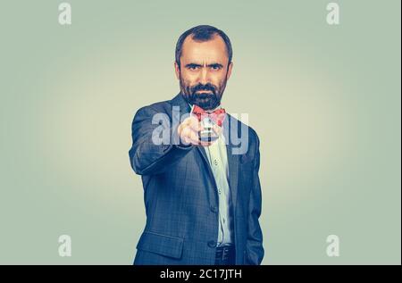Adult displeased bearded man in elegant suit with red bow tie holding remote tv control. Mixed race bearded model isolated on green yellow uniform gra Stock Photo