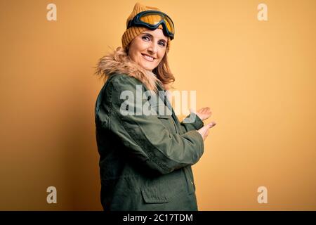 Middle age beautiful blonde skier woman wearing snow sportwear and ski goggles Inviting to enter smiling natural with open hand Stock Photo