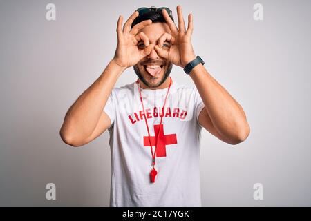 Young handsome lifeguard man with beard wearing t-shirt with red cross and whistle doing ok gesture like binoculars sticking tongue out, eyes looking Stock Photo