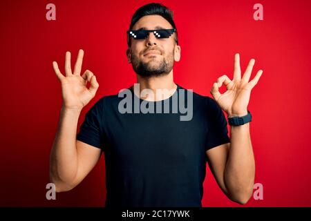 Young handsome man wearing funny thug life sunglasses over isolated red background relax and smiling with eyes closed doing meditation gesture with fi Stock Photo