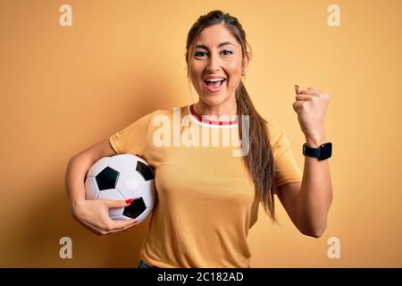 Beautiful brunette player woman playing soccer using football ball over yellow background screaming proud and celebrating victory and success very exc Stock Photo
