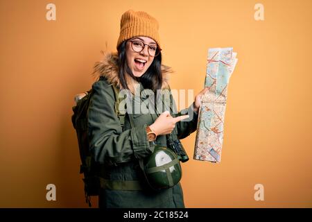 Young hiker woman wearing hiking backpack looking at tourist map over yellow background very happy pointing with hand and finger Stock Photo