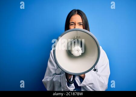 Hispanic doctor woman wearing medical white coat shouting angry on protest through megaphone. Yelling excited on ludspeaker talking and screaming news Stock Photo