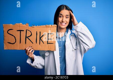 Young doctor woman wearing stethoscope holding cardboard banner protesting in strike stressed with hand on head, shocked with shame and surprise face, Stock Photo