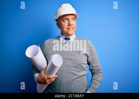 Middle age handsome grey-haired architect man wearing safety helmet holding blueprints smiling looking to the side and staring away thinking. Stock Photo