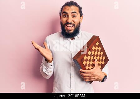 Young arab man holding chess celebrating achievement with happy smile and winner expression with raised hand Stock Photo