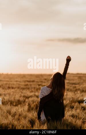 Young beautiful girl in white dress and jacket walking through the wheat orange field on a sunny summer day. Feel freedom with arm stretched to the Stock Photo