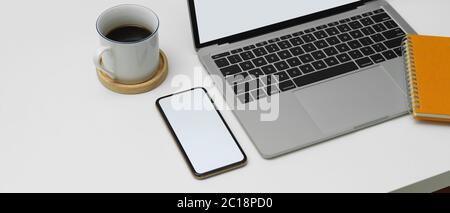 Cropped shot of modern worktable with blank screen smartphone, laptop and coffee cup on white desk Stock Photo