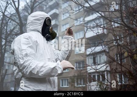 Man in gas mask and protective clothing monitoring city environment. Scientist checking tree leaves, taking piece using tweezers for analysis. Environmental disaster, smoky empty atmosphere in yard. Stock Photo