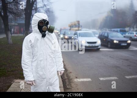 Close up of scientist looking at road with cars, waiting for green light. Selective focus of man in gas mask and protective costume posing near pedestrian crossing. Environmental disaster, fog. Stock Photo