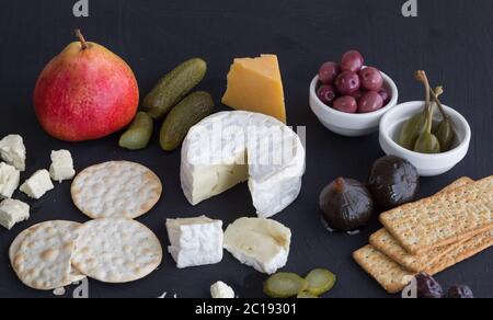 Camembert cheese, pear, gherkins, cheddar cheese, olives, figs and crackers on black background Stock Photo