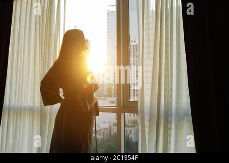 young girl in bathrobe opens window transparent curtains in apartment side view Stock Photo