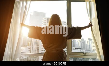 girl silhouette in bathrobe opens window transparent curtains looking at buildings outside in hotel room backside view Stock Photo