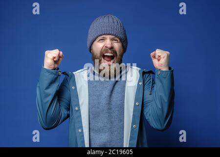 Man in a knitted hat and waterproof jacket is happy that he is going on a travel, makes a gesture with hands and screams joyfully. Stock Photo