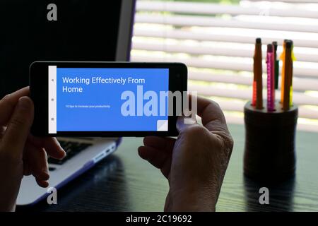 Person reading google slides presentation on android mobile phone in a home office with laptop in background. Text Working Effectively from Home Stock Photo