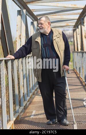 Caucasian senior man with walking stick standing on an metal bridge over small river Sura in Ukraine Stock Photo