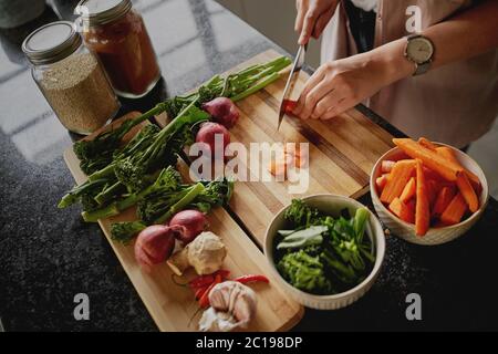High-angle closeup of young female hands chopping fresh orange carrot on board while in modern kitchen Stock Photo