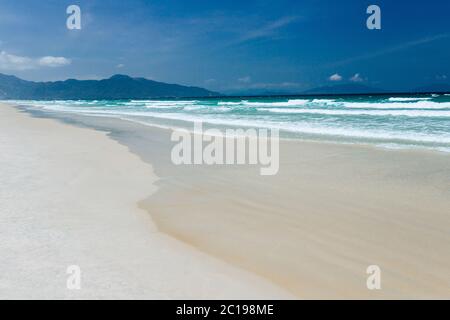 Landscape view of the sandy sea beach. Calm waves, foam. Blue sky with white clouds. An empty long beach with ocean and mountain views in the backgrou Stock Photo