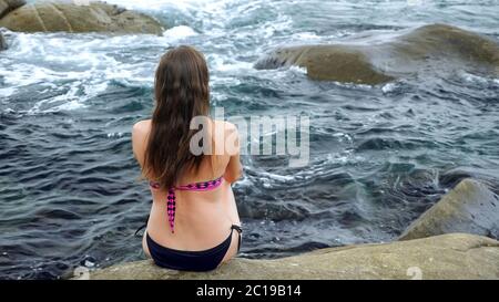 long dark haired slim woman in stylish bikini sits on large old rock washed by deep blue ocean waves backside view Stock Photo