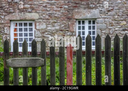 A pretty stone cottage with stone lintels and white mullioned windows stands surrounded by grass and a picket fence. Stock Photo