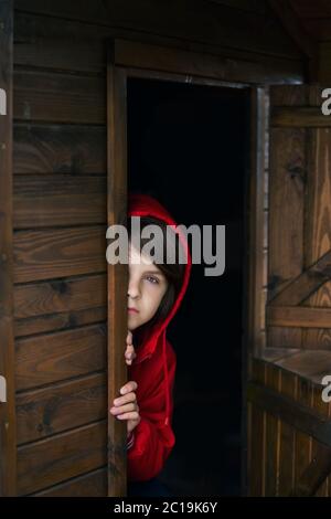 Preteen boy in red sweatshirt, hiding behind a wooden door, looking scared and sad Stock Photo