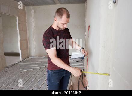 Side view of serious man in shirt holding notebook and using measuring tape in kitchen with underfloor heating pipes. Handyman taking measurement while working on renovation of apartment.
