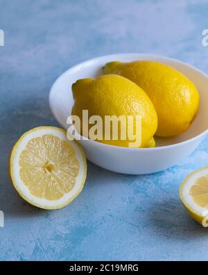 Whole lemons in a white bowl with sliced lemon against blue textured background Stock Photo