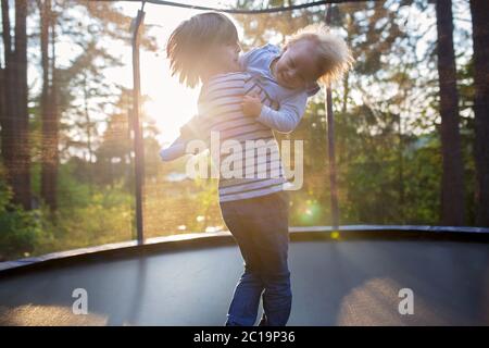 Sweet preteen boy and his toddler brother jumping on trampoline. Happy children jumping on sunset, making different shapes Stock Photo