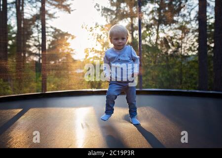 Sweet toddler baby boy jumping on trampoline. Happy little blond child jumping on sunset for the first time and having fun outdoor Stock Photo