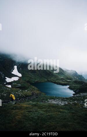 Hiker in a yellow jacket walking in a foggy mountains near the glacier Capra lake,Romania Stock Photo