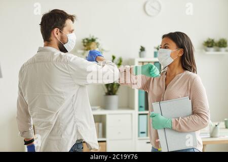 Waist up portrait of two colleagues wearing face masks bumping elbows while greeting each other at work in office Stock Photo