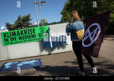 15 June 2020, Hamburg: An activist of the environmental protection movement Extinction Rebellion (XR) stands with a flag in front of the entrance to the Norddeutscher Rundfunk (NDR) during a protest, on whose wall a banner with the inscription 'Climate crisis in the media! Shows the extent of the ecological crisis' is hanging on the wall. With a blockade in front of the entrance to the NDR premises in Hamburg-Lokstedt, environmental activists have called on the station to replace the term climate change with climate crisis in its reporting. Photo: Christian Charisius/dpa Stock Photo