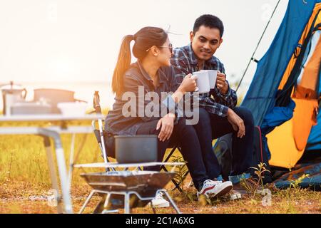 couple sitting and drinking coffee to happily near the lake at outdoors camping. Lifestyle, holiday, long weeked and camping concept. Stock Photo