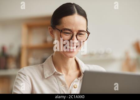 Warm-toned portrait of young businesswoman wearing glasses smiling happily while looking at laptop screen, copy space Stock Photo