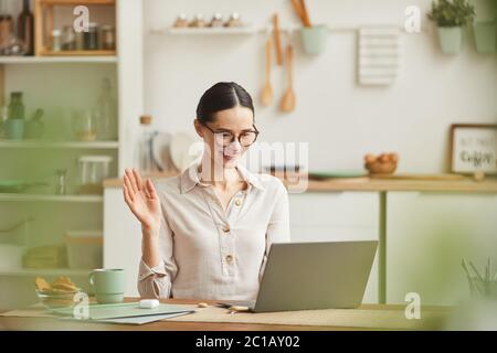 Warm-toned portrait of smiling young woman waving at camera during online meeting while working from home, copy space Stock Photo