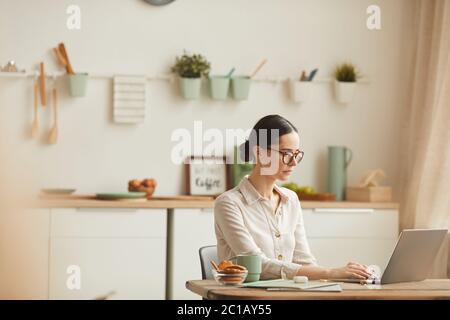 Warm-toned side view portrait of elegant young woman wearing glasses while using laptop at cozy home office workplace, copy space Stock Photo