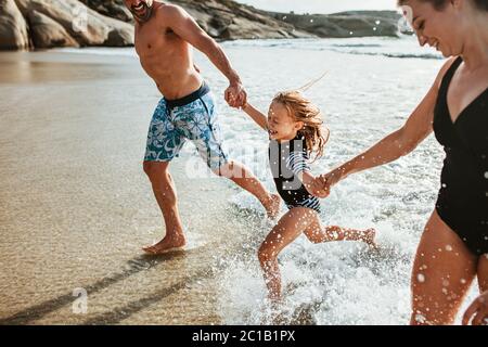 Parents holding hands of their daughter and running out of the sea water at the beach. Family enjoying a summer weekend on the beach.