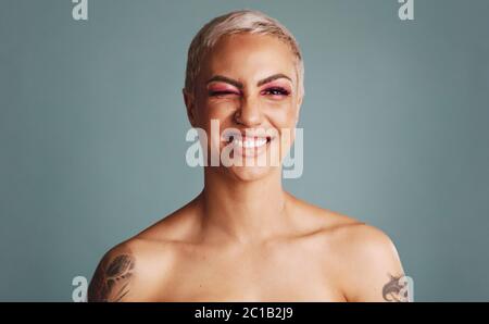 Smiling woman looking at camera and winking an eye. Beautiful female model with blonde pixie hairstyle having tattoo on her hands standing on grey bac Stock Photo
