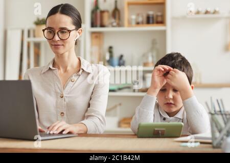 Warm toned portrait of young mother working at home office with son using smartphone beside her in cozy kitchen interior Stock Photo