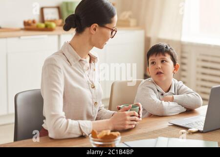 Warm-toned portrait of young mother talking to son while doing homework together sitting at table in cozy kitchen interior, copy space Stock Photo