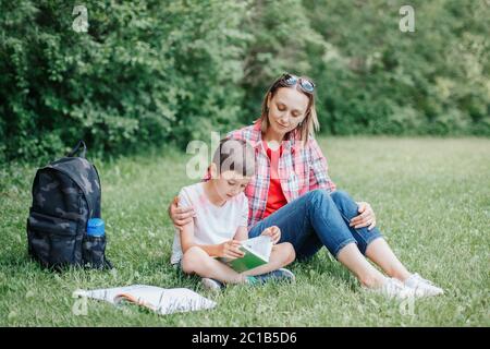 Young Caucasian mother reading book with son together. Family doing school homework in park outdoor. Mom helping child kid with education learning Stock Photo