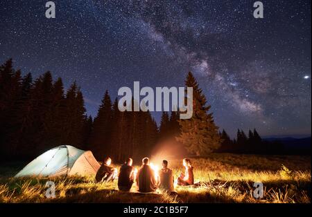 Night summer camping in the mountains, spruce forest on background, sky with stars and milky way. Back view group of five tourists having a rest together around campfire, enjoying fresh air near tent. Stock Photo