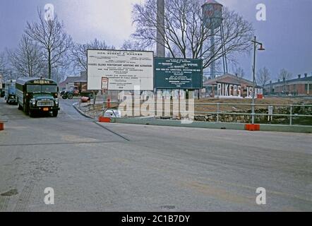 The road up to the Rikers Island prison complex on a winter's day, East River, New York, USA 1963. The large 3-D letters spelling out 'Rikers Island' are on the right, in front of the water tower. A prison bus is departing (left). The billboards indicate major building works are being carried out under NY Mayor Robert F Wagner. Stock Photo