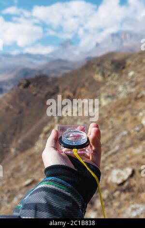 A man's hand holds a pocket magnetic compass for navigation against the background of a rocky slope and epic rocks under a blue Stock Photo